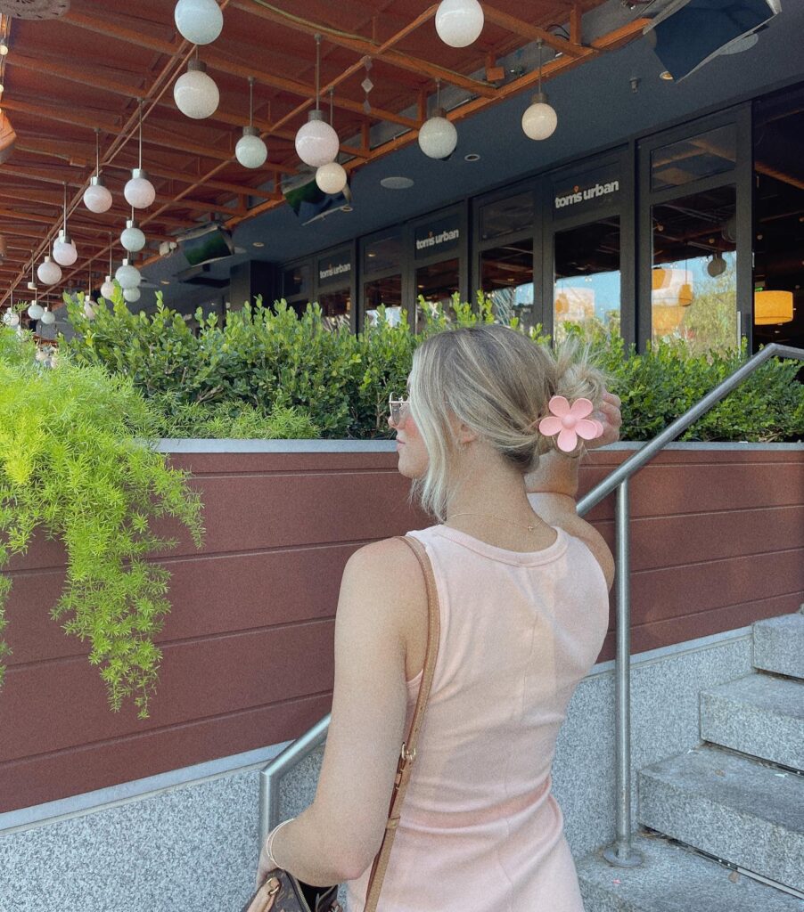 A woman viewed from behind, with blonde hair gathered into a casual low updo and secured with a large, pink flower claw clip.