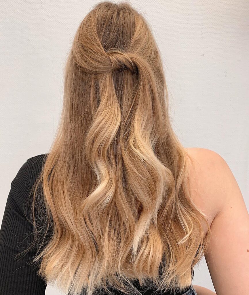 A woman with long wavy honey blonde hair styled in a simple twisted half up knot with loose waves cascading down her back.