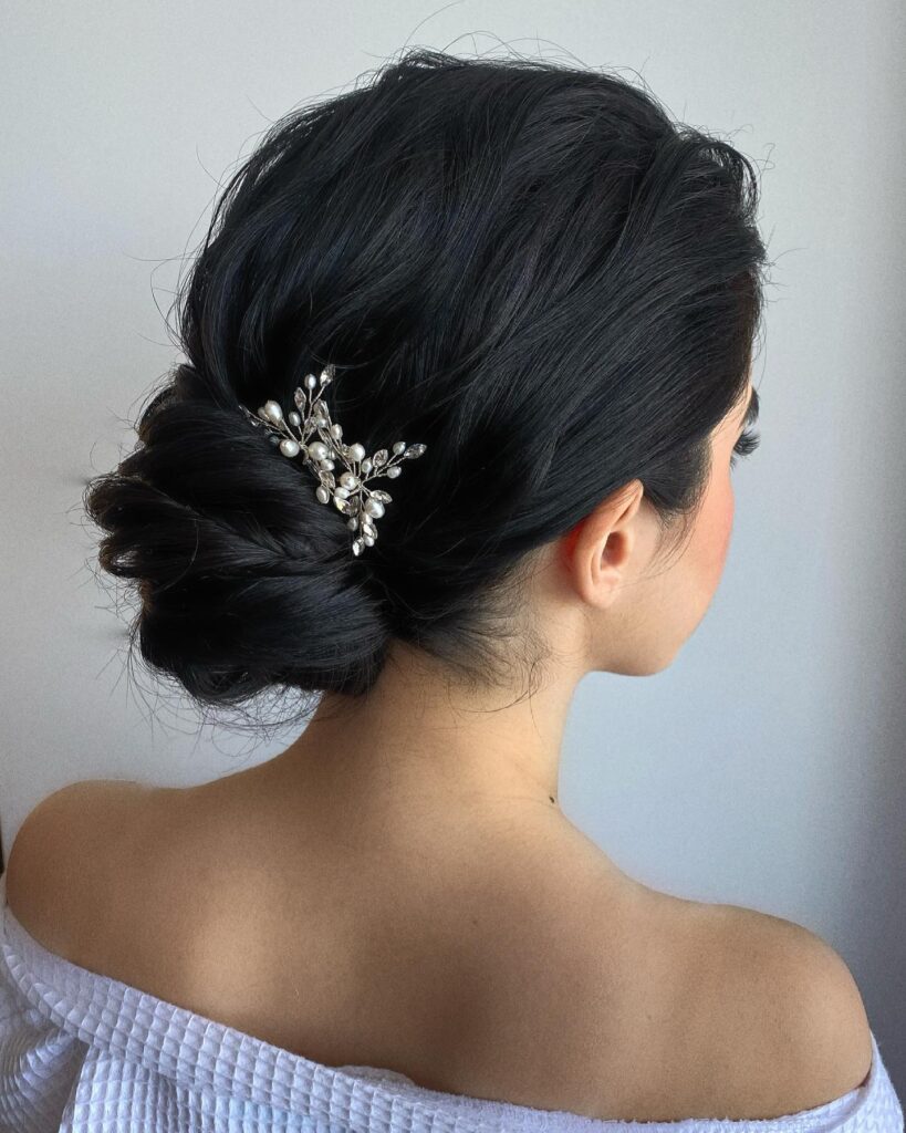 A woman with long black hair styled into a textured low chignon bun secured with a pearl and crystal leaf hair accessory.