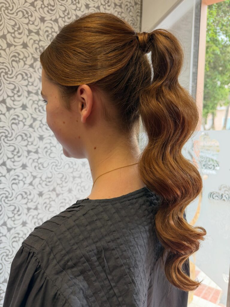 Side view of a woman with long auburn hair styled in a high wrapped ponytail with smooth defined glam waves.