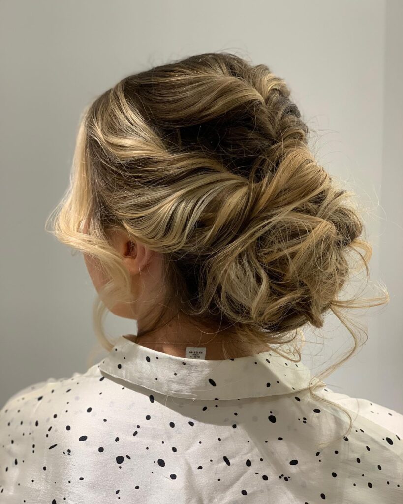 A woman with highlighted blonde hair styled in a loose braided messy low bun with soft face framing strands.