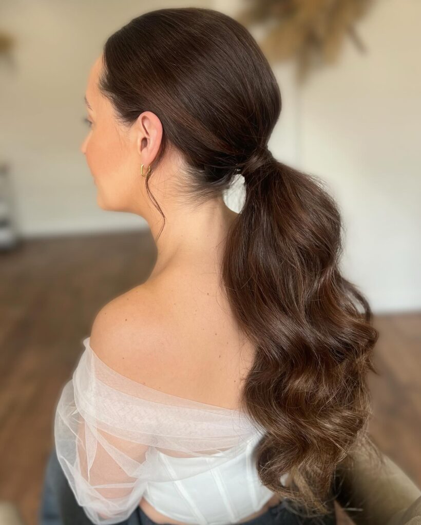 Side profile of a woman with long dark brown hair styled in a sleek wrapped low ponytail with smooth soft waves.