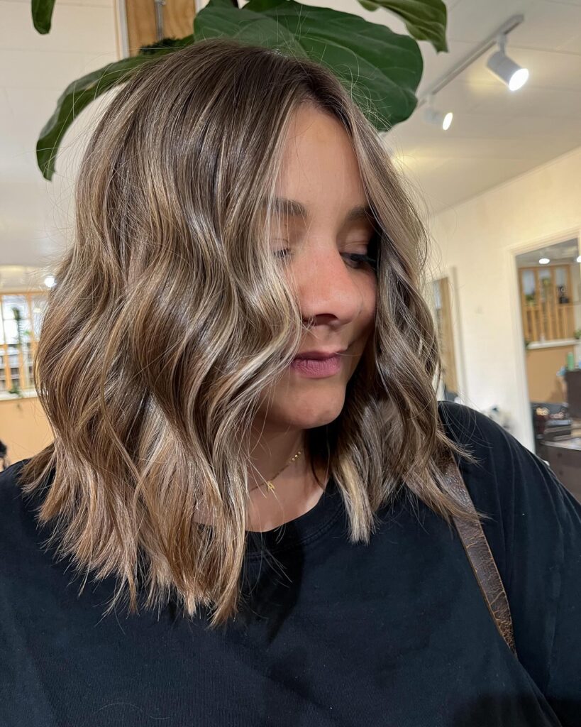 A woman with a wavy shoulder length lob featuring sandy blonde highlights and textured ends.