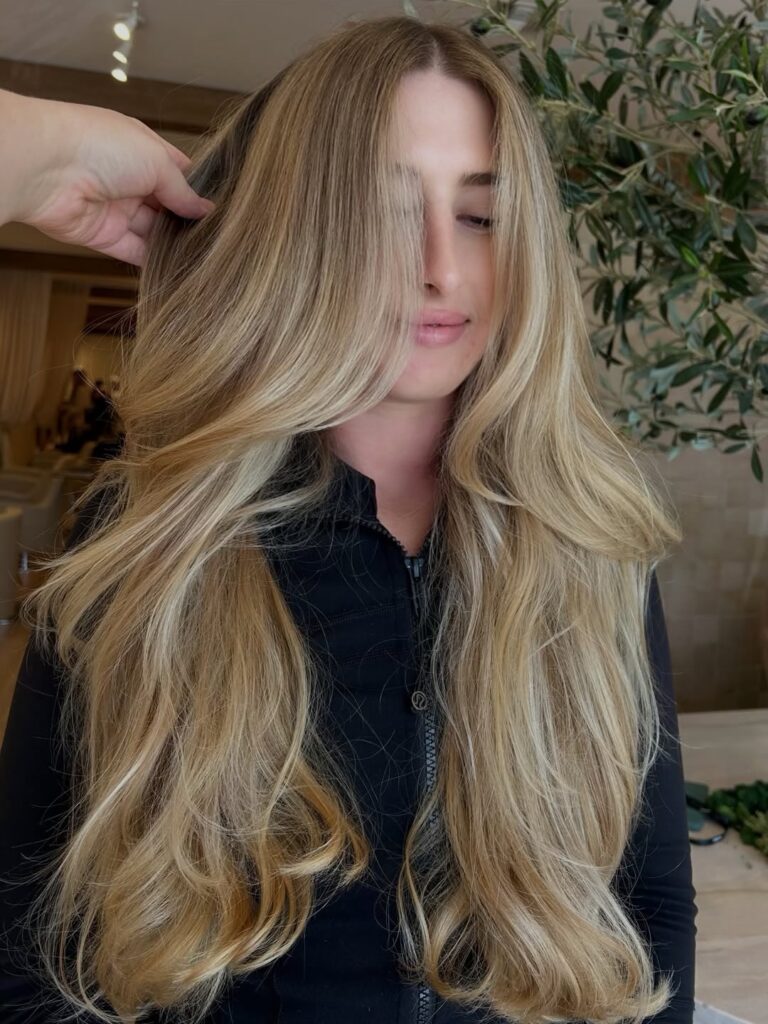 A woman with long, voluminous, golden blonde layered hair is shown after a beautiful blowout.