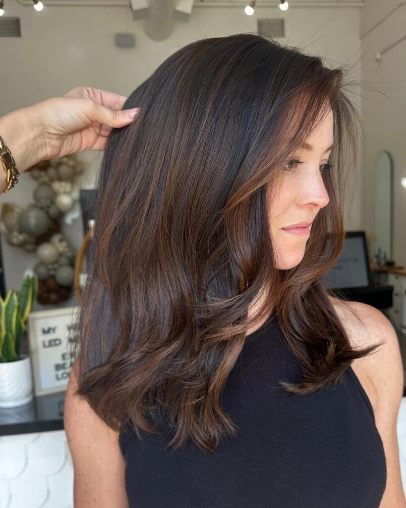 A woman with medium length dark chocolate brown hair styled in soft waves with subtle chestnut highlights.