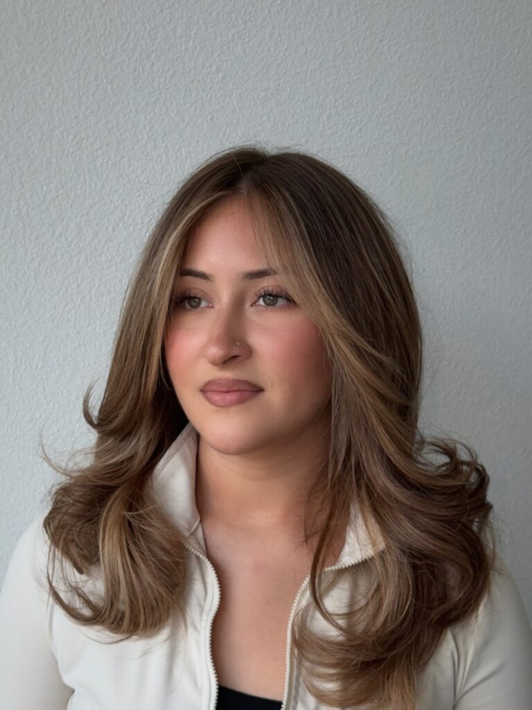 A young woman with a luminous light brown layered haircut features face-framing curtain bangs and subtle blonde balayage highlights styled in soft, bouncy waves.