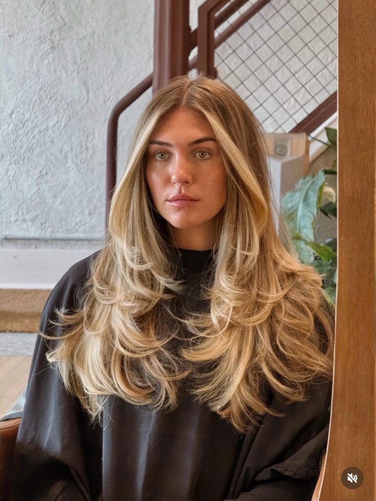 A woman with long layered golden blonde hair features a bright platinum money piece and flipped bouncy ends.
