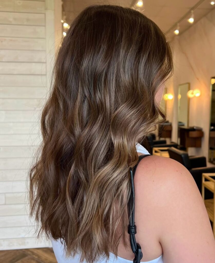 A woman with long dark brown hair shows off soft waves featuring warm chestnut and caramel highlights.