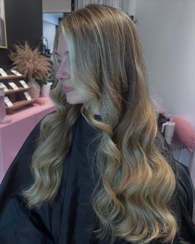 A portrait of a woman showing off her long honey blonde balayage styled in loose dimensional waves.