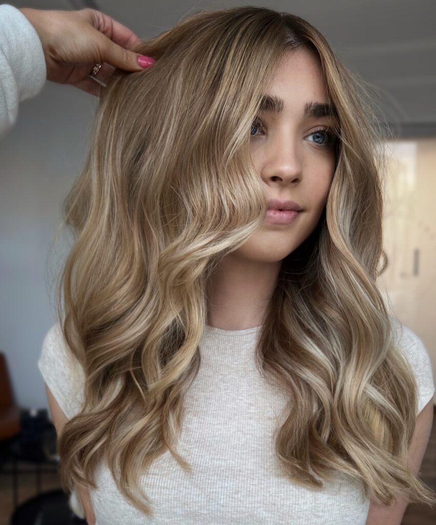 A young woman with medium long golden honey blonde hair styled in loose beach waves with bright face framing highlights.