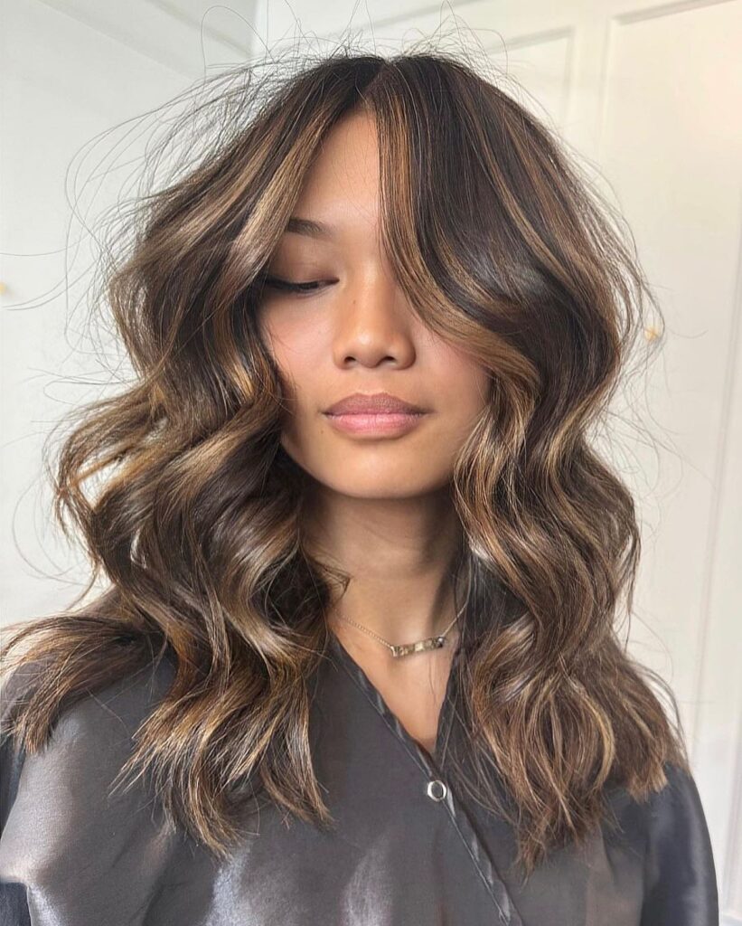 A woman with medium length dark brown hair showcases beachy waves adorned with warm caramel highlights.