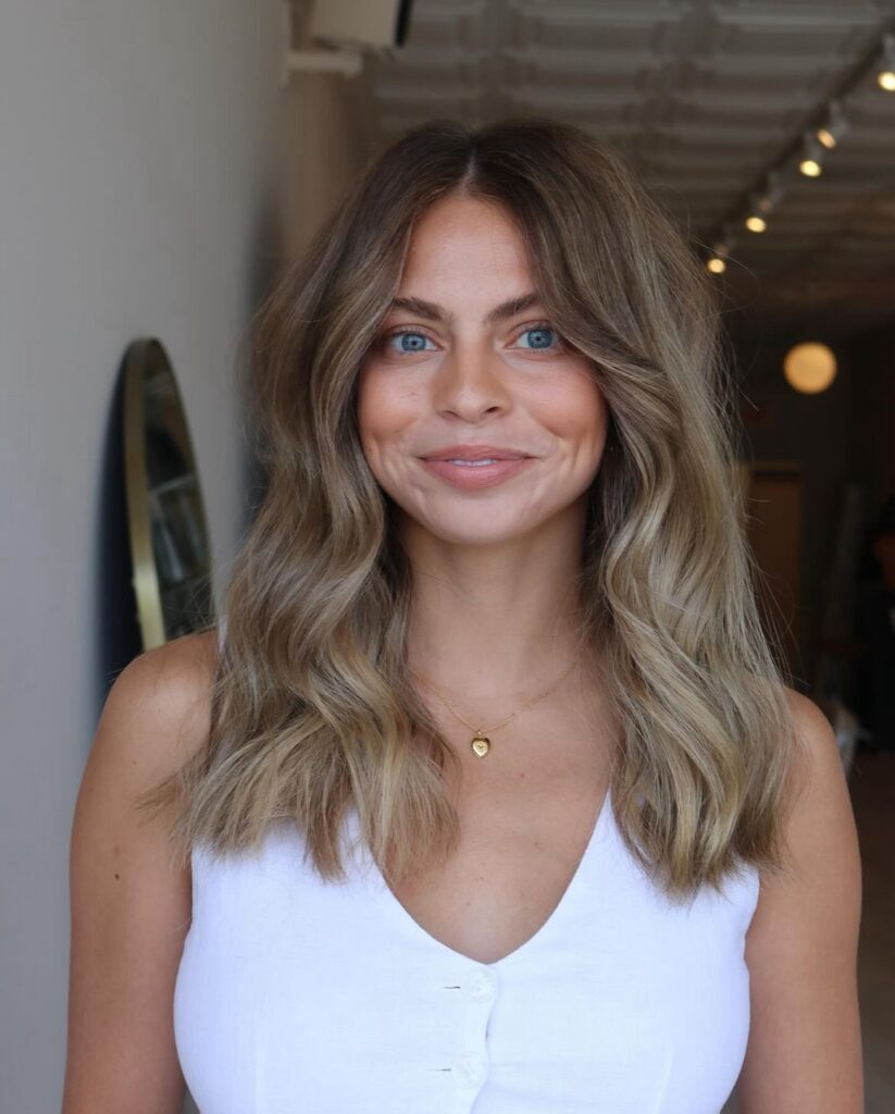 A smiling woman with shoulder length sandy ash blonde hair styled in soft waves with curtain bangs and dark roots.
