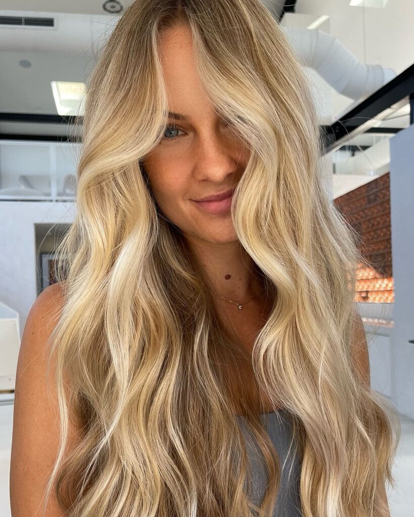 A smiling woman with long bright creamy blonde hair styled in soft loose beach waves with face framing highlights.