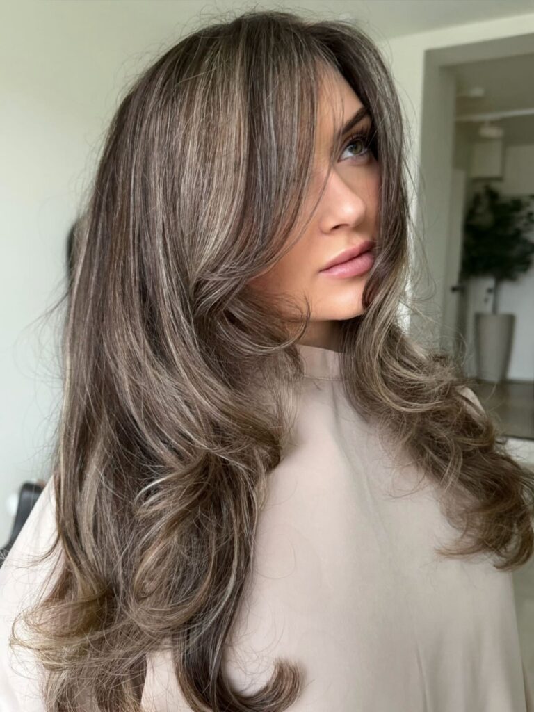 A woman with long cool ash brown hair showcases a bouncy blowout featuring fine blonde highlights and face framing curtain bangs.