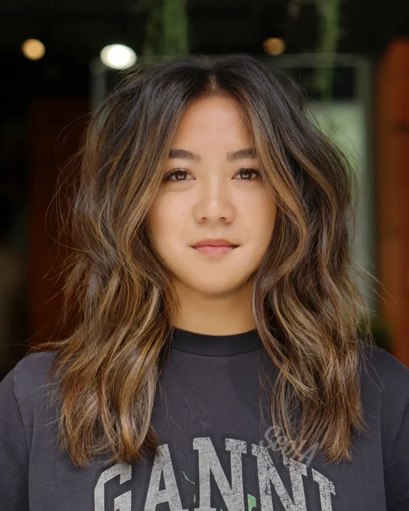 A portrait of a woman showing off a textured shoulder length brown bob styled with subtle caramel highlights and soft wavy definition.