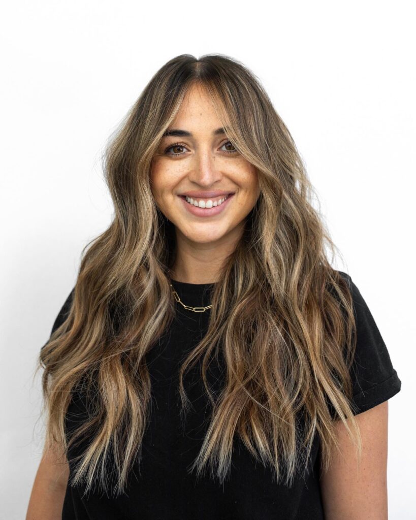A smiling woman with long wavy dark brown hair featuring soft caramel balayage highlights and center parted curtain bangs.