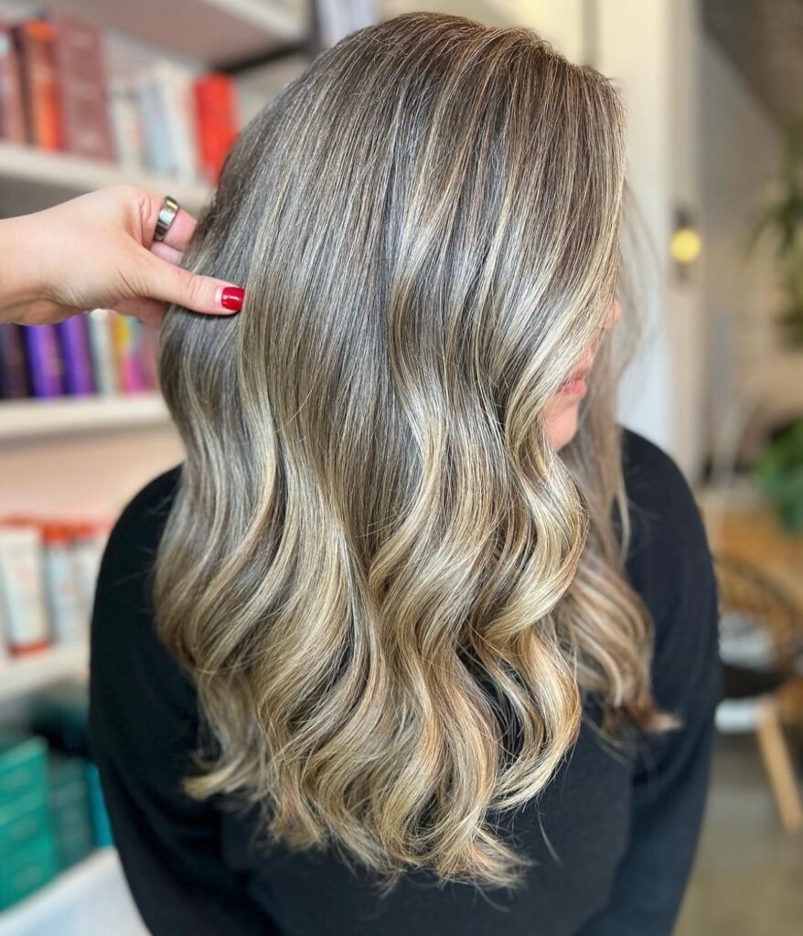 A woman with long, beautiful hair styled with soft, voluminous waves is showing off her ultra glossy silver blonde balayage highlights that blend with a light brown root.
