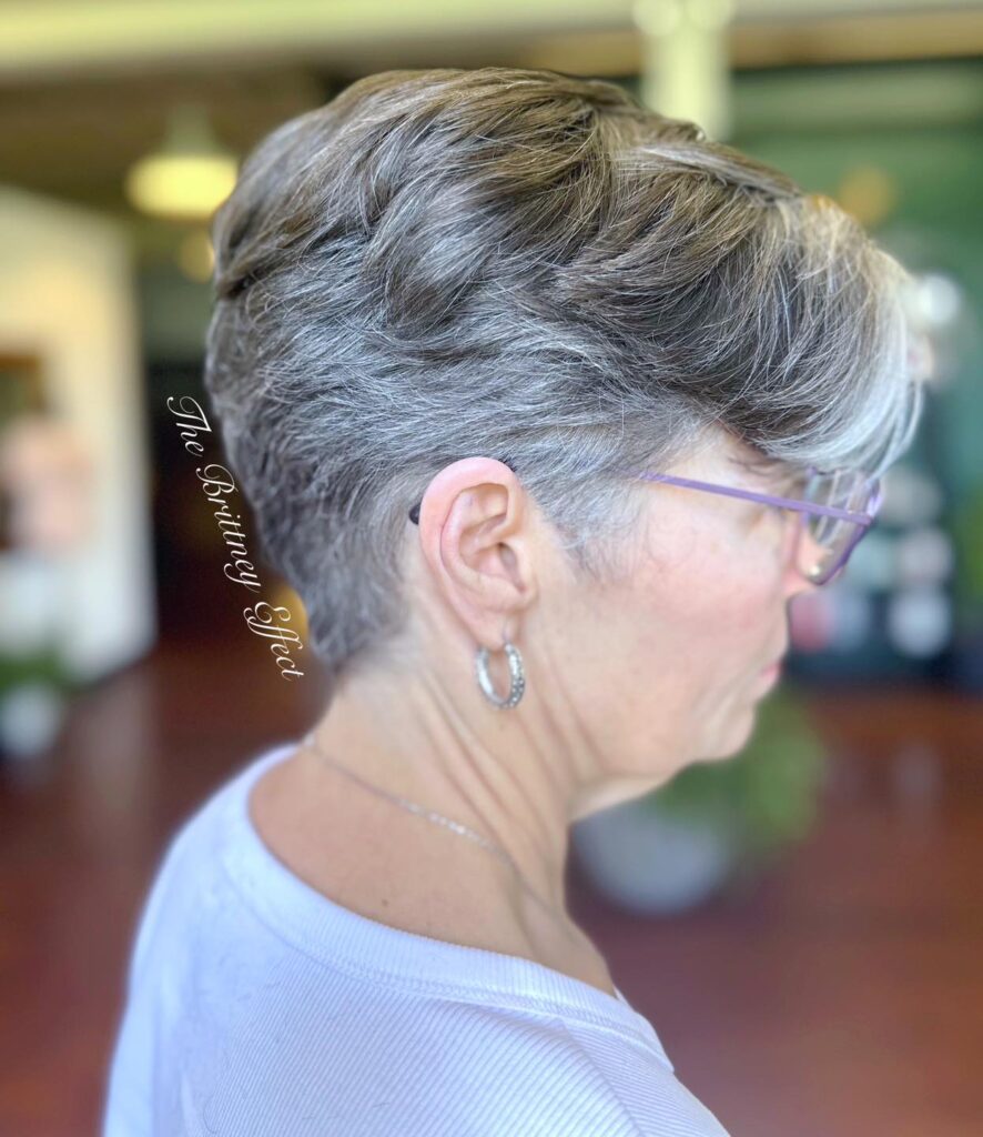 A side profile of a woman with glasses showcasing a short charcoal pixie haircut with textured silver waves on top.