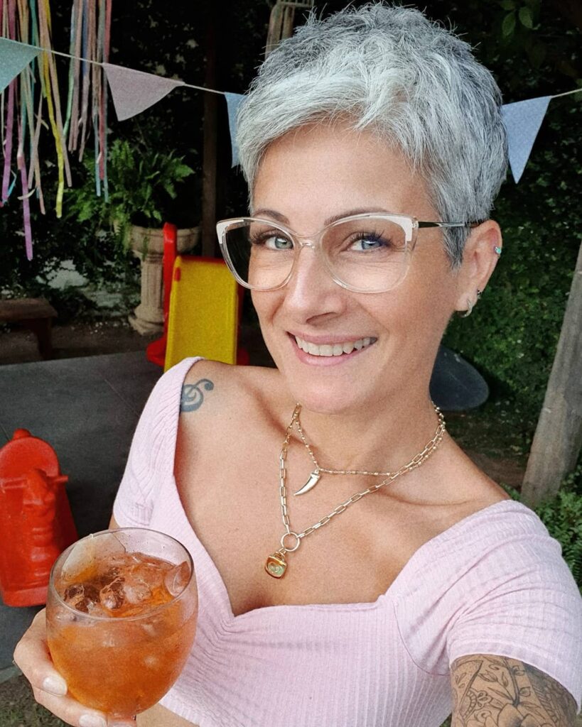 A woman with a spiky bright icy silver pixie cut and clear glasses smiles while holding a glass.