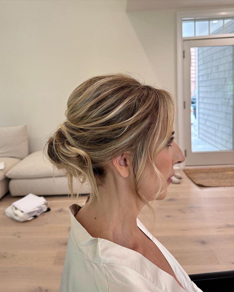 A side profile of a woman wearing an elegant low chignon updo with honey blonde highlights and face framing tendrils.