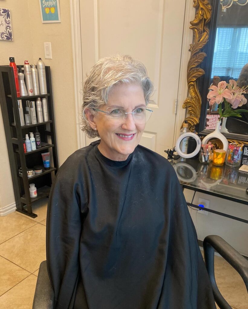 A smiling woman with glasses showcasing an ear length feathered silver pixie bob haircut with soft waves at a salon.