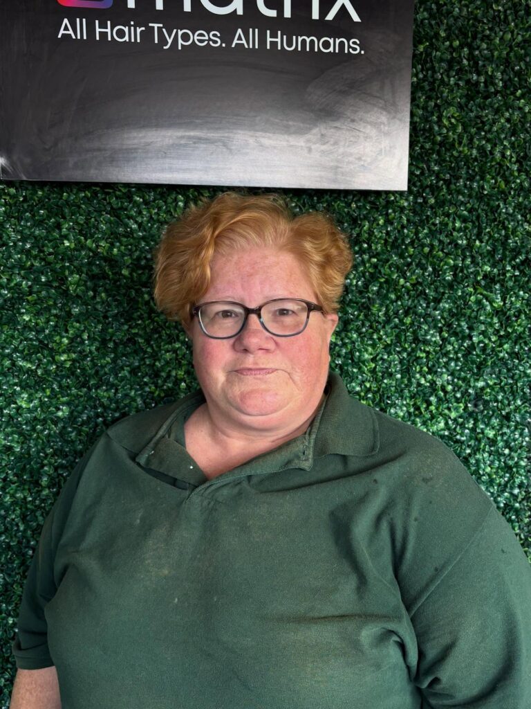 A woman with glasses posing in front of a green wall showcasing a short golden copper pixie haircut with soft voluminous waves.
