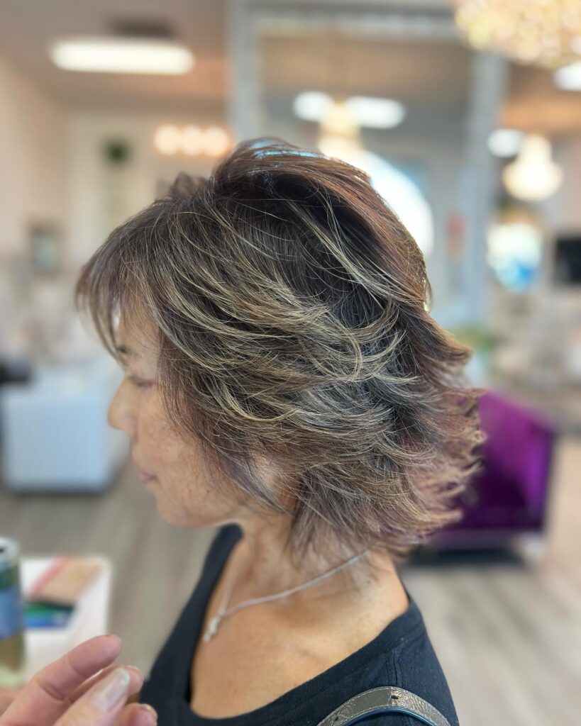 A woman viewed from the side profile in a salon showcases a neck length bronde shag haircut with wispy feathered layers and fine blonde highlights.