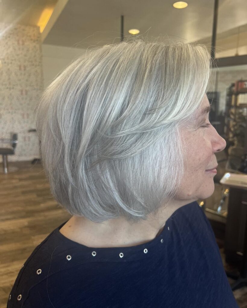 A woman viewed from the side profile in a salon, showcasing a sleek chin length silver white bob haircut with long feathered layers.