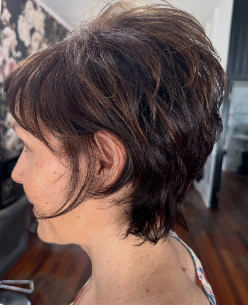 A woman viewed from the side profile showcases a short textured brunette pixie shag haircut with warm honey highlights and wispy layers.