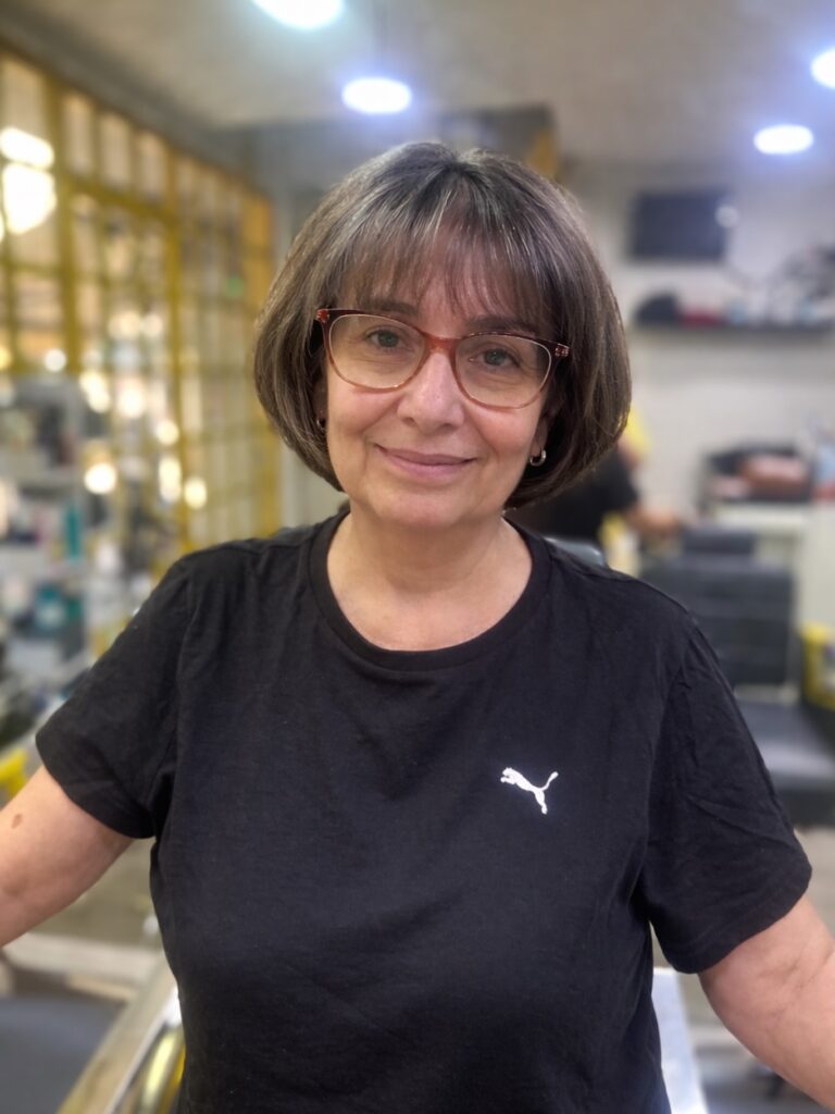 A smiling older woman wearing glasses and a black t-shirt showcases a classic chin length ash brunette bob with wispy bangs.