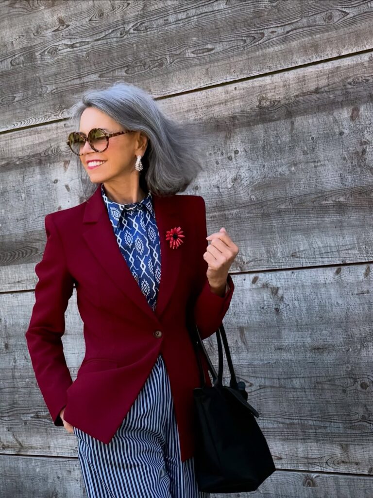 A woman with a wavy charcoal grey bob wearing a red blazer and sunglasses standing against a wooden wall.