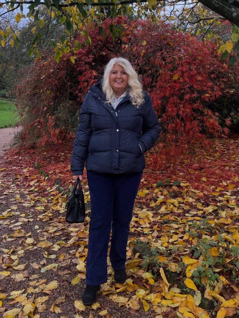 A smiling woman over 60 posing in front of red autumn leaves showcases long platinum blonde hair styled in soft bouncy waves.
