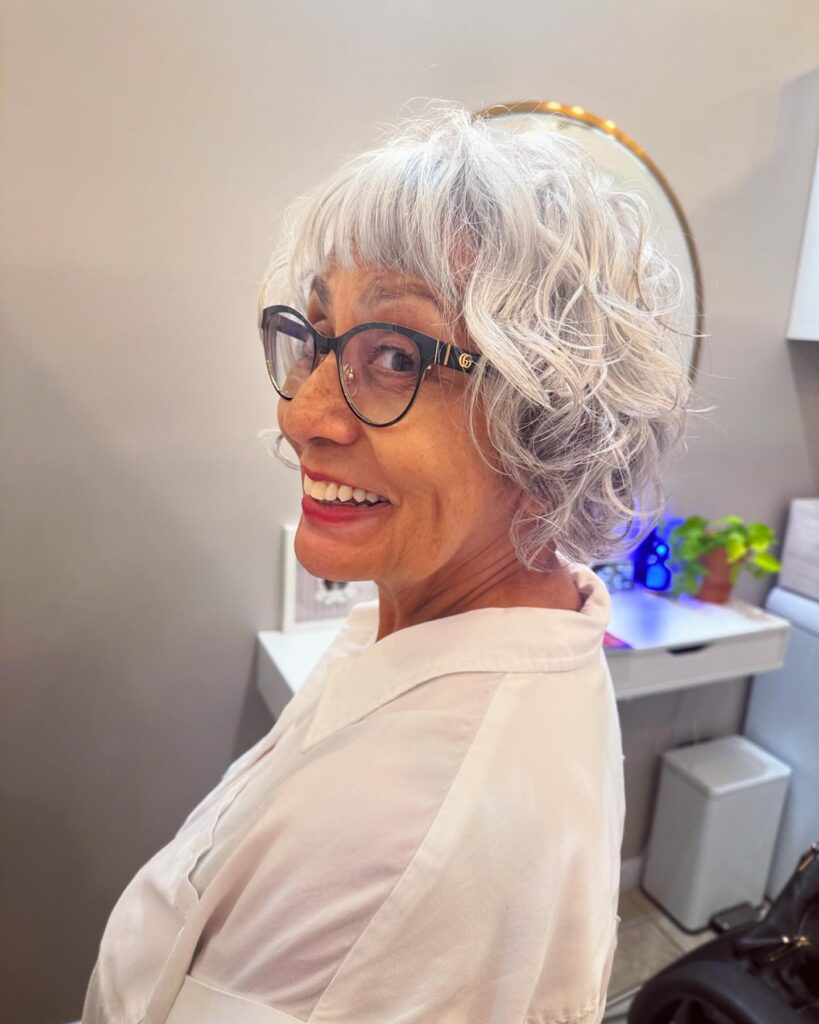 A smiling woman with an ear length icy silver curly pixie bob and wispy bangs.
