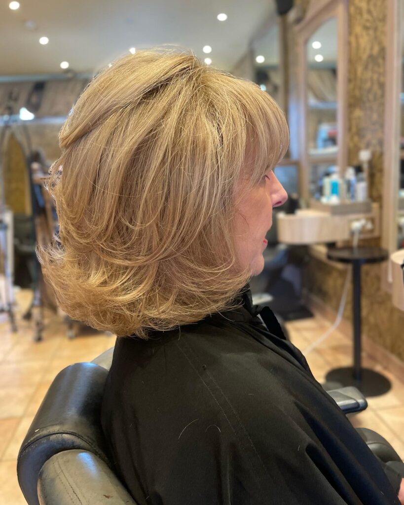 A side profile view of a woman in a salon chair showcasing a neck-length layered caramel blonde bob haircut with full bangs and a rounded bouncy style.