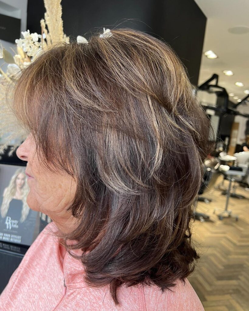 A side profile view of a woman with a layered brunette shag haircut featuring bright caramel highlights and feathered volume.