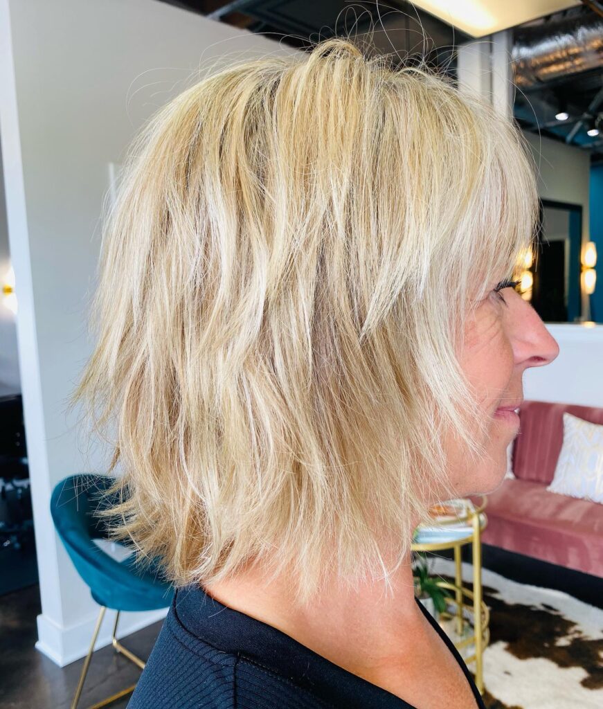 A side profile view of a woman with a choppy, bright blonde shaggy lob haircut featuring razored layers and soft bangs.