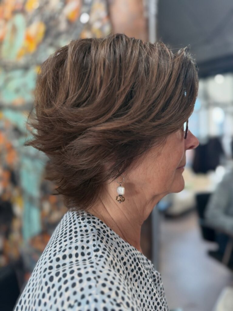 A side profile view of a woman with a texturized short salt and pepper shag haircut featuring choppy layers and soft bangs.