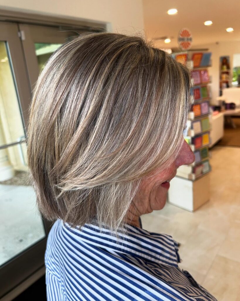 A side profile view of a woman with a collarbone-length ash blonde lob haircut featuring feathered, face-framing layers.