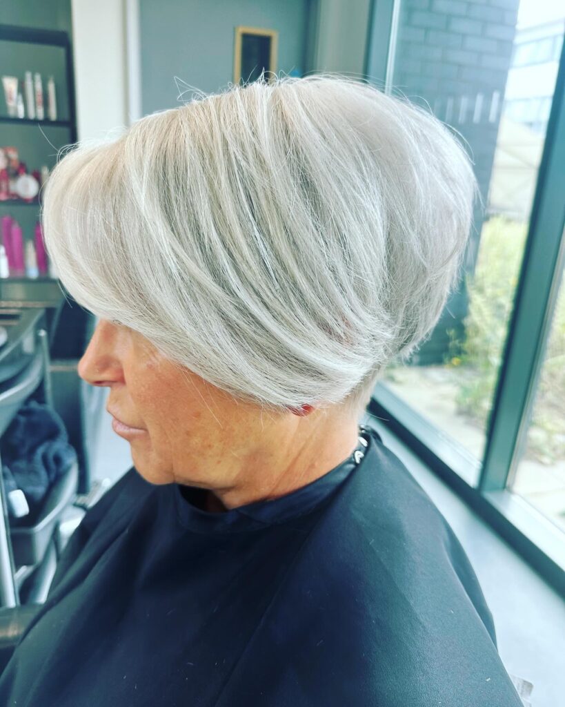 A side profile view of a woman with a sleek, angled silver bob haircut featuring long side layers and a stacked back.