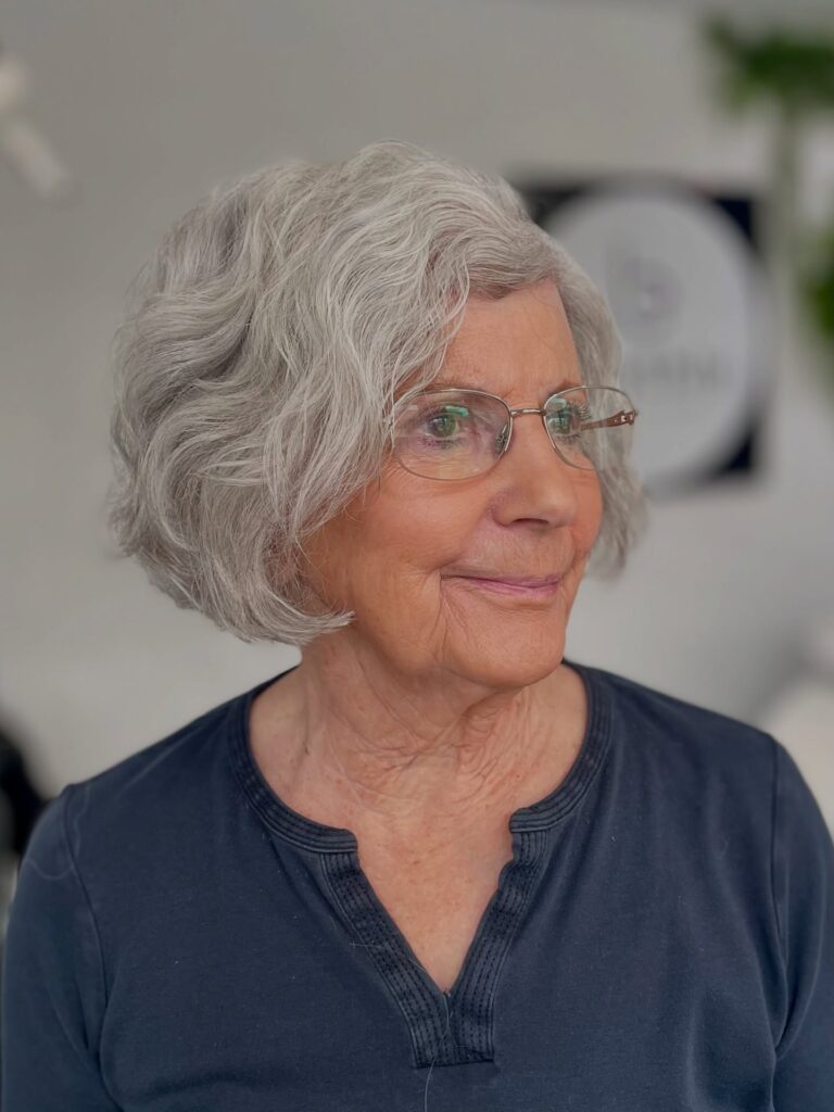 An older woman wearing glasses smiles while showcasing a textured chin-length silver bob with soft waves and side-swept bangs.