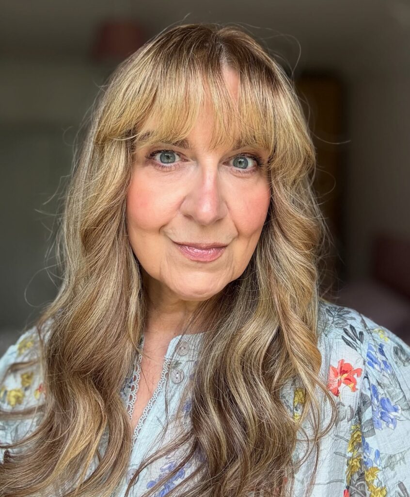 A woman over 60 with long wavy honey blonde hair and light wispy bangs smiling at the camera.