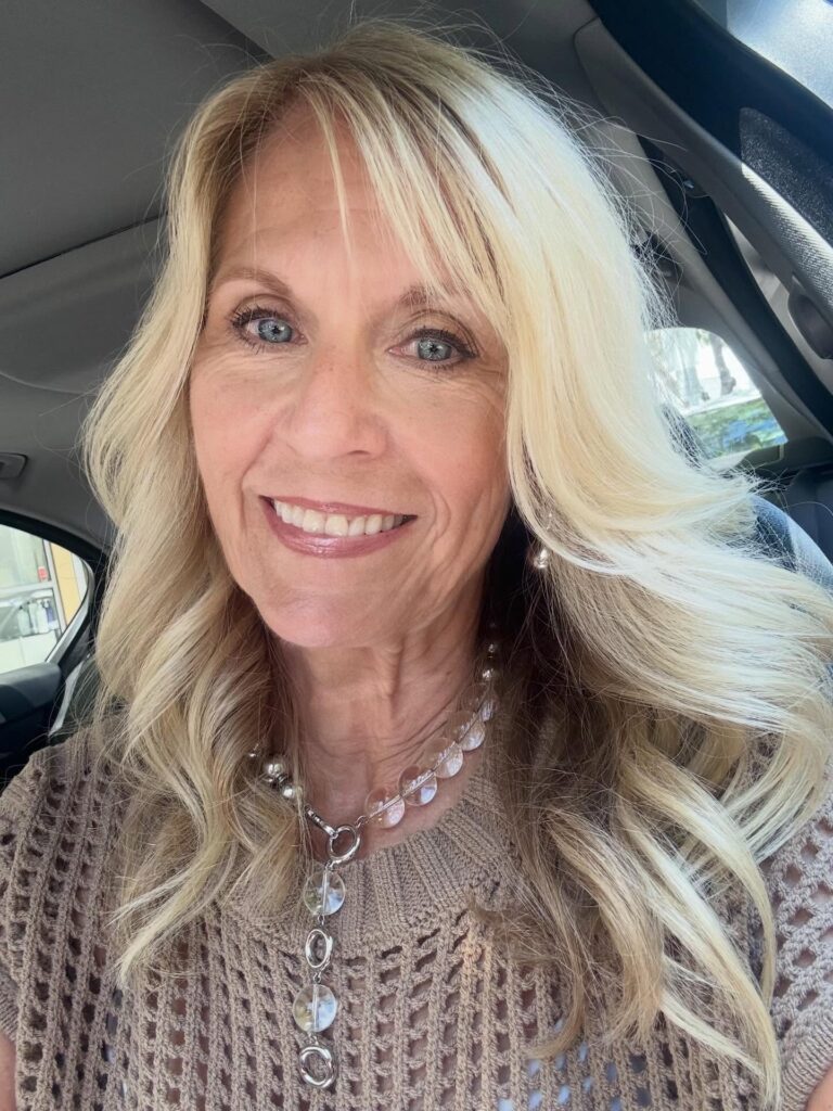 A smiling woman over 60 with long blonde hair styled in soft voluminous waves with side-swept bangs.