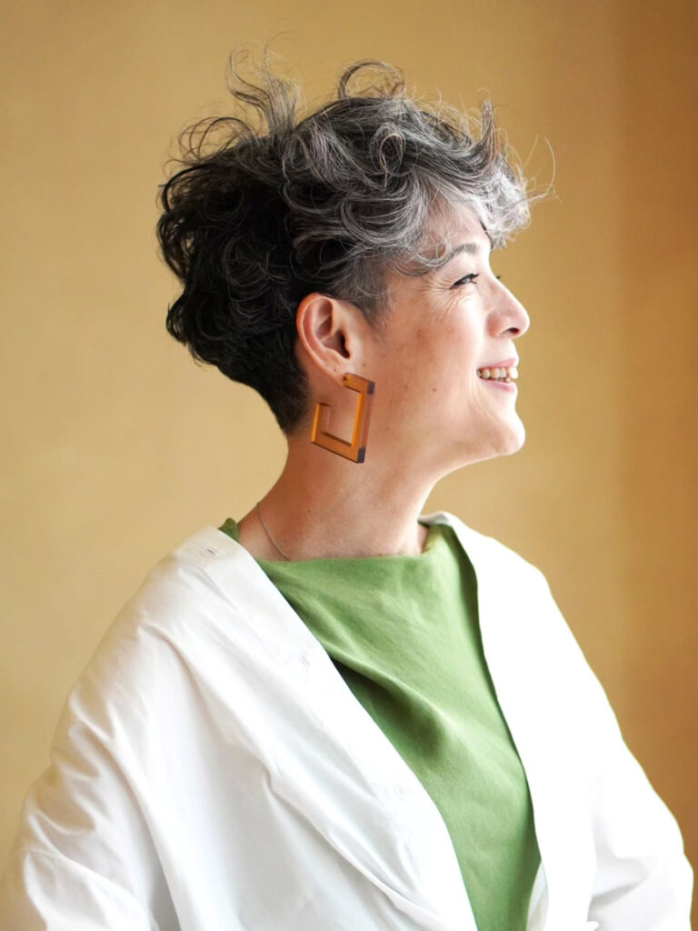 A woman over 60 with a high-volume salt-and-pepper curly haircut featuring closely cropped, tapered sides, smiling in profile.