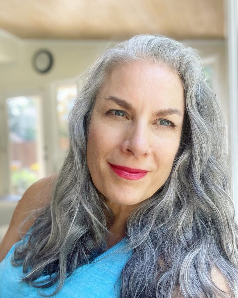 A close-up photograph of a woman with long wavy silver and dark charcoal hair looking directly at the camera.