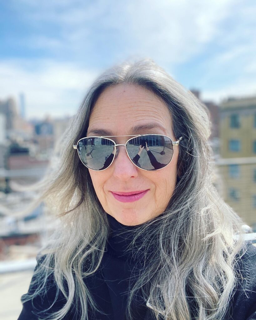 A woman wearing aviator sunglasses and a black turtleneck shows off her long wavy silver hair with a city skyline background.