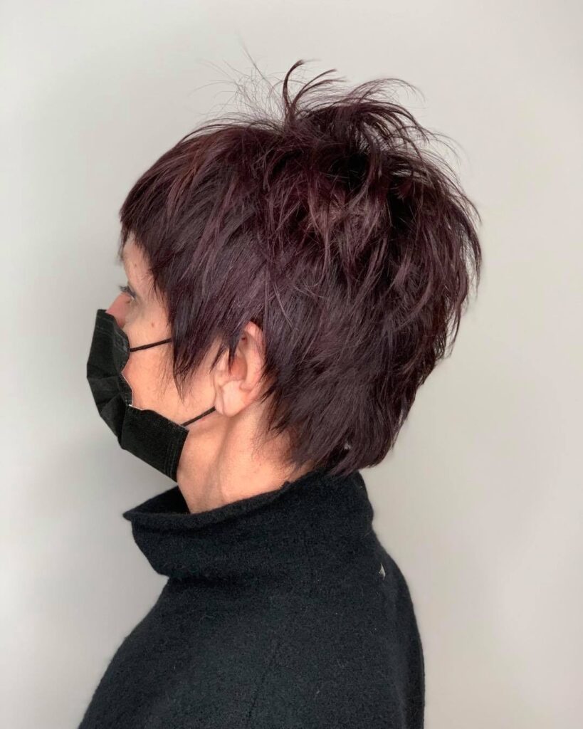 A woman showing a side profile view of a messy plum-colored shag pixie haircut with piecey top layers.
