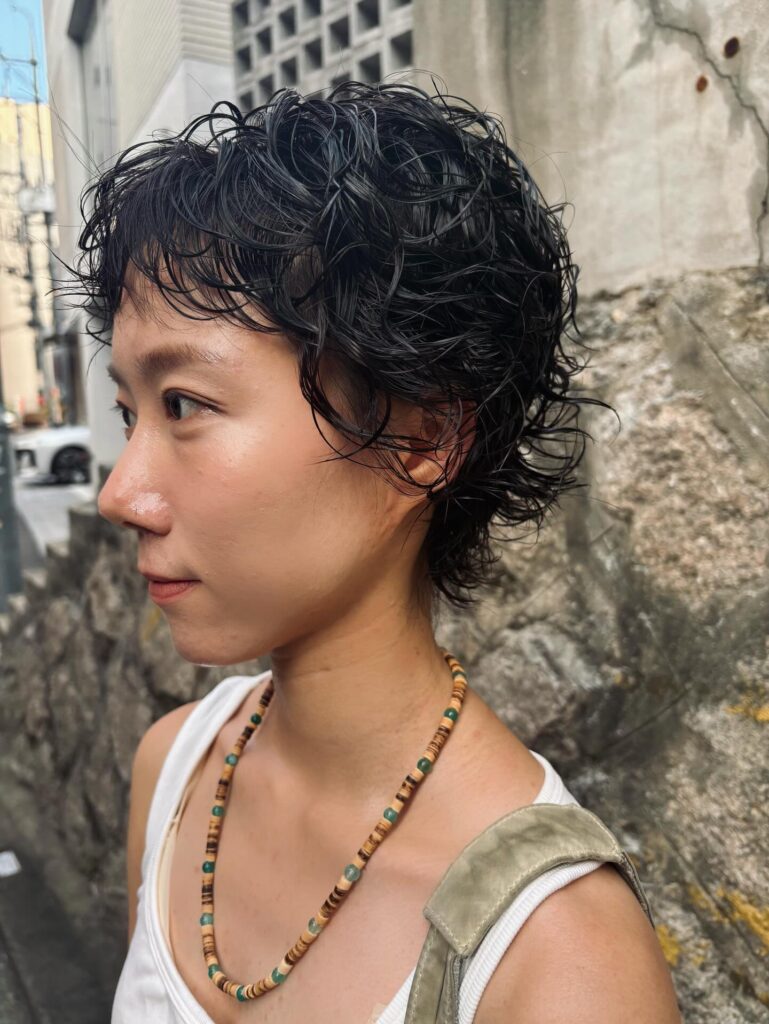 A woman showing a profile view of a wet look jet black curly pixie haircut with wispy micro bangs.