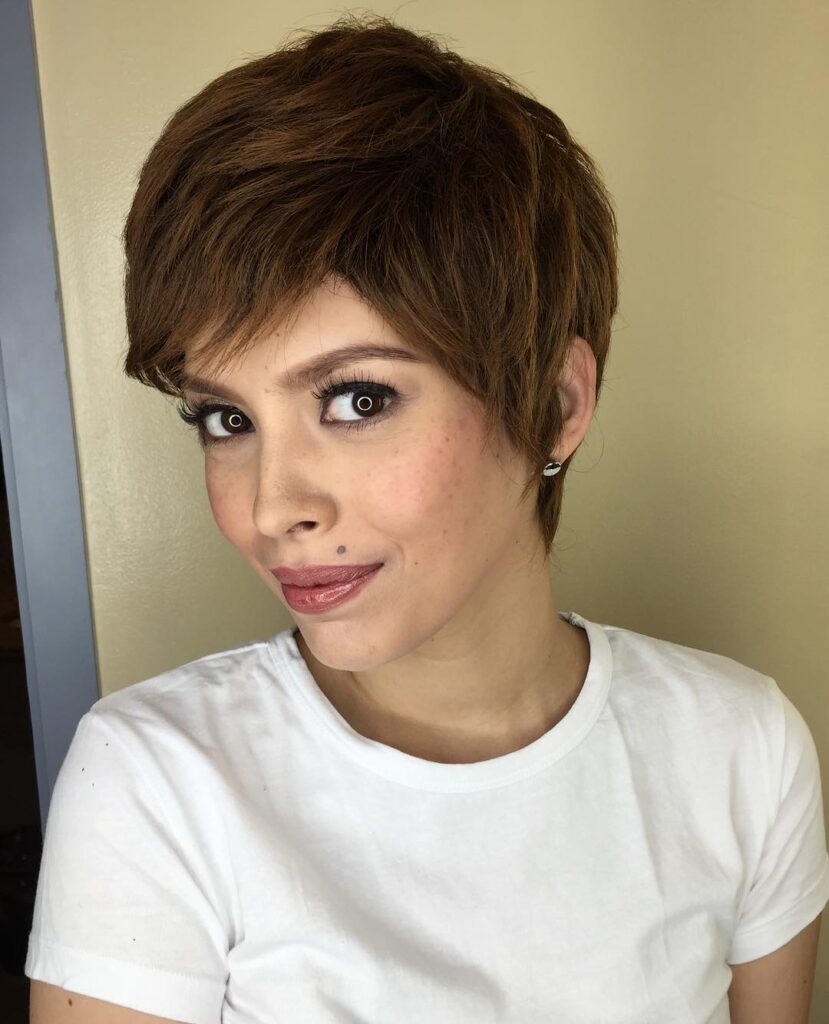 A woman with a warm chestnut brown layered pixie haircut and soft side bangs looking forward at the camera.