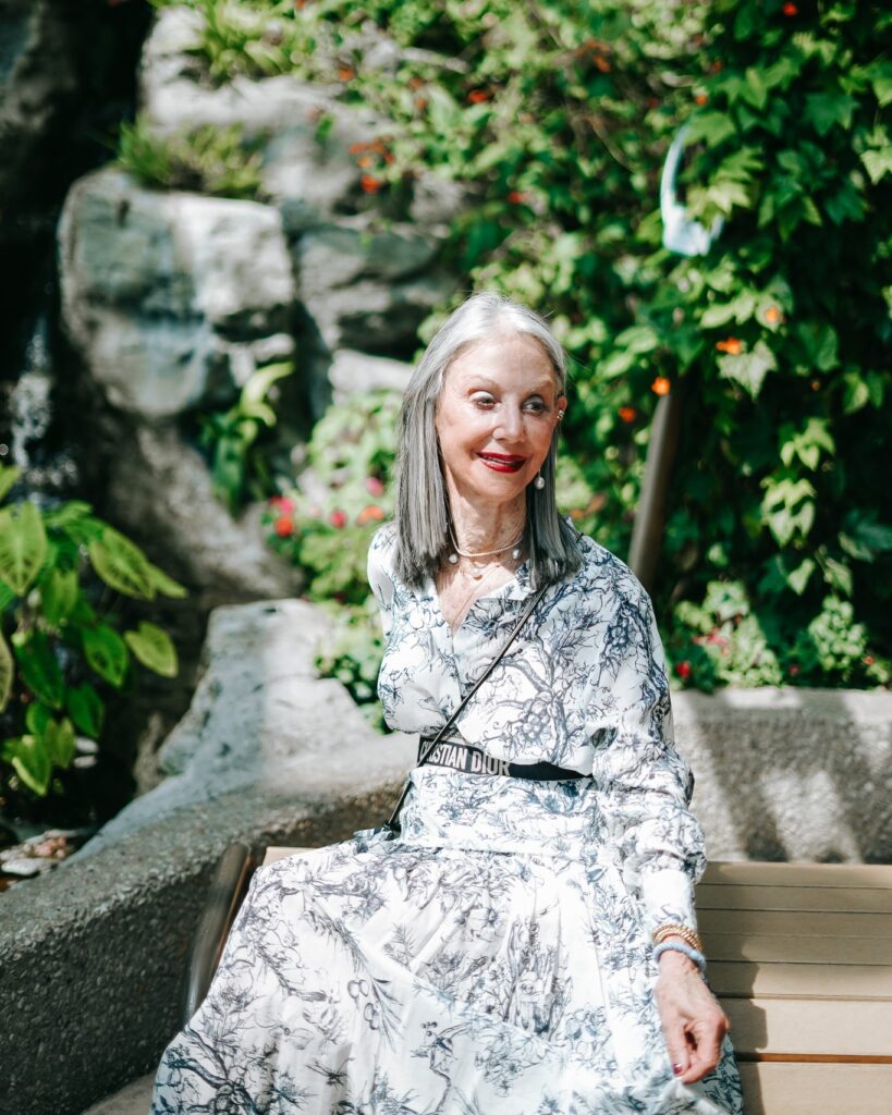 A woman over 60 sitting on a bench with a straight salt and pepper lob hairstyle parted down the middle.