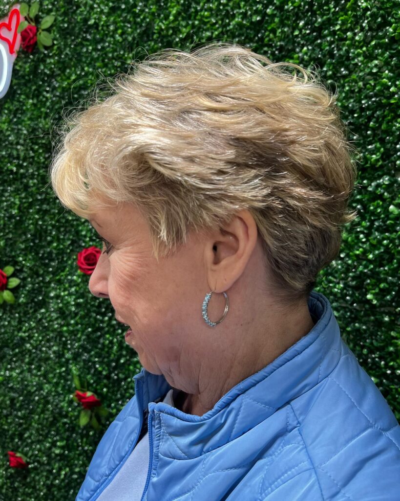 A side profile view of a woman over 60 with a short tapered honey blonde pixie hairstyle featuring wavy layers on top.
